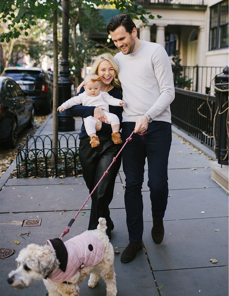 Familia caminando por la calle con su bebé en brazos y su perro con abrigo rosa, sesión familiar en exterior